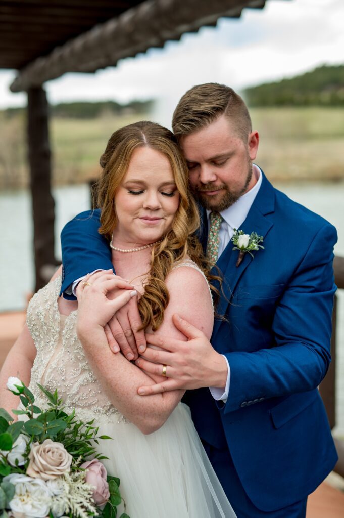 The groom holds his bride tightly in a tender embrace, showcasing their deep connection and love. They stand against a beautiful backdrop, with the bride's gown flowing elegantly, capturing a moment of pure emotion and intimacy between the couple.