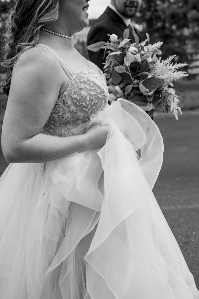 The bride gracefully holds onto the delicate fabric of her wedding dress as she walks, her gown flowing behind her. The moment captures the elegance and anticipation of her journey to the ceremony, set against a serene backdrop at Spruce Mountain Ranch.