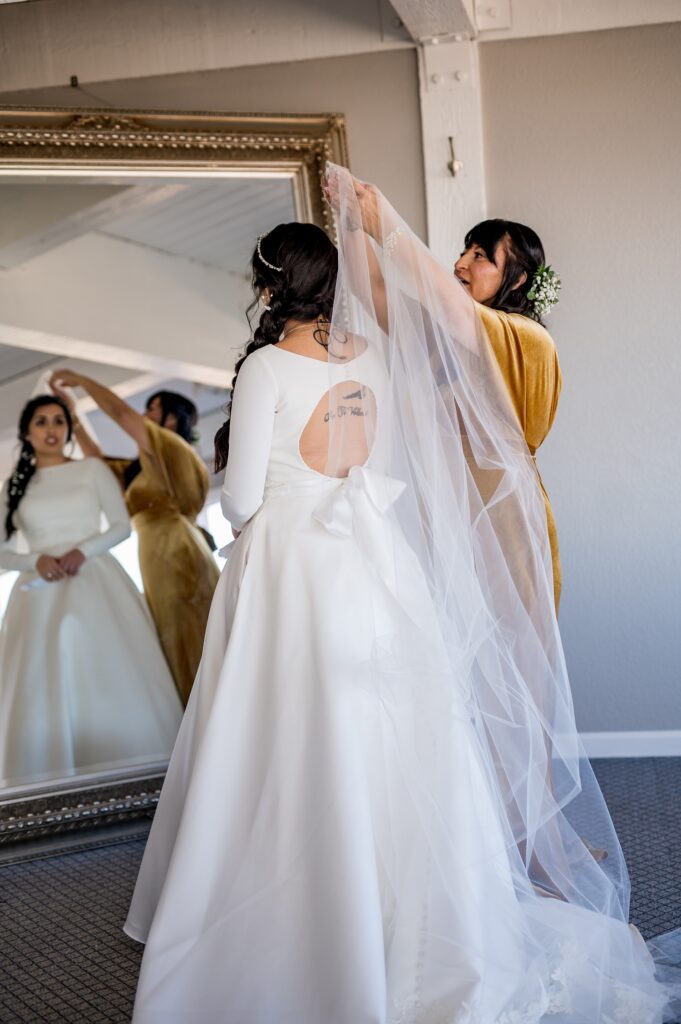 The mother of the bride gently places the veil on her daughter, carefully adjusting it as they share a tender, heartfelt moment. The bride smiles softly, and warm light illuminates the scene, highlighting the love and joy between them on this special day.