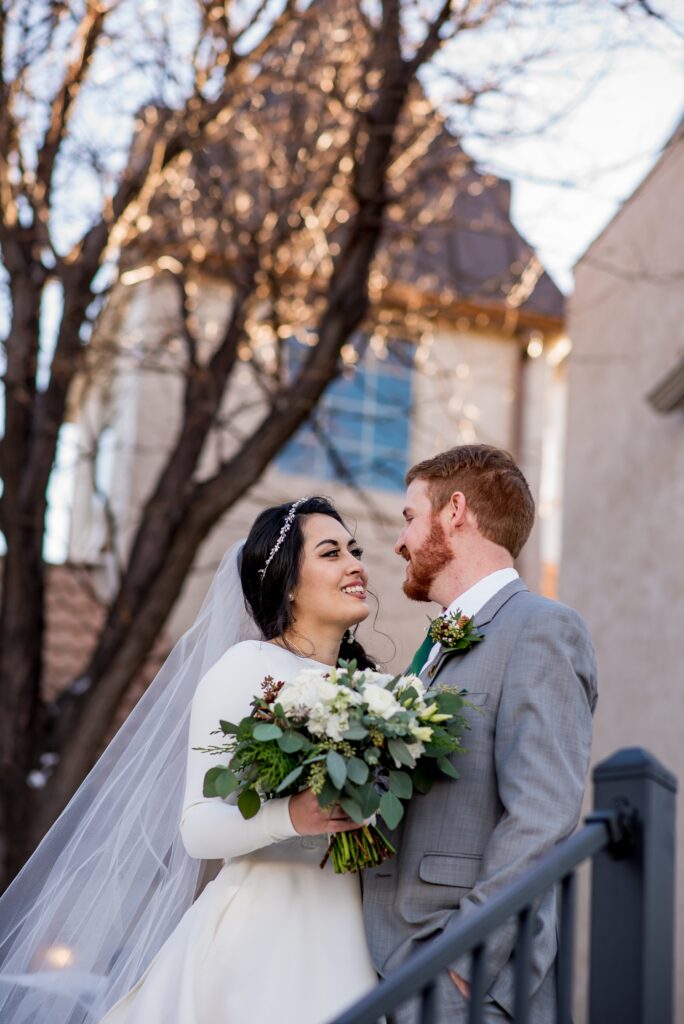 A Colorado wedding couple gazes lovingly into each other’s eyes before their ceremony at Brittany Hill. Surrounded by the breathtaking views of the Denver skyline and mountains, they share an intimate moment filled with anticipation and joy, capturing the romance of their wedding day.