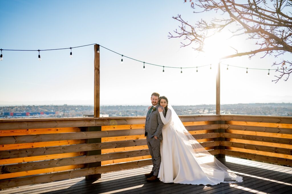 The bride and groom stand on the back deck of Brittany Hill during sunset, silhouetted by the warm glow of the sky. With the stunning Colorado mountains and Denver skyline in the background, they share a quiet, romantic moment, capturing the breathtaking beauty of their winter wedding day.