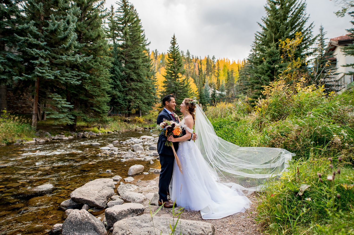 A bride and groom embrace by a peaceful mountain stream surrounded by vibrant fall foliage and towering evergreens at the Sonnenalp Hotel in Vail, Colorado. The bride’s flowing veil and romantic bouquet complement the serene fall setting, perfectly captured by a Vail wedding photographer.