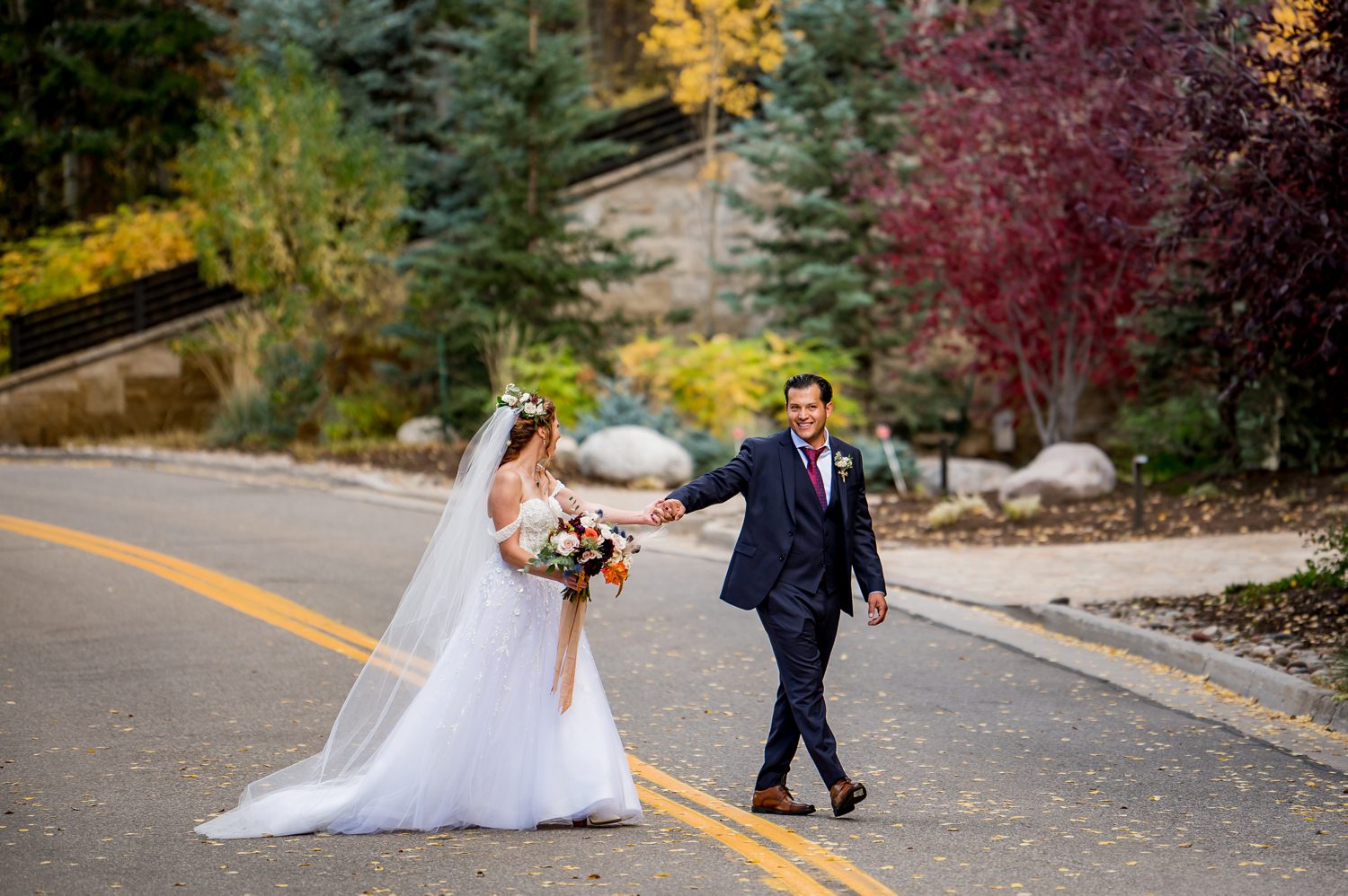 A bride and groom walking hand-in-hand on a scenic mountain road in Vail, Colorado, surrounded by vibrant fall foliage, golden aspens, and lush evergreens. The bride carries a bouquet of autumn florals while her flowing gown and veil add romantic elegance to the scene, captured by a Vail wedding photographer.