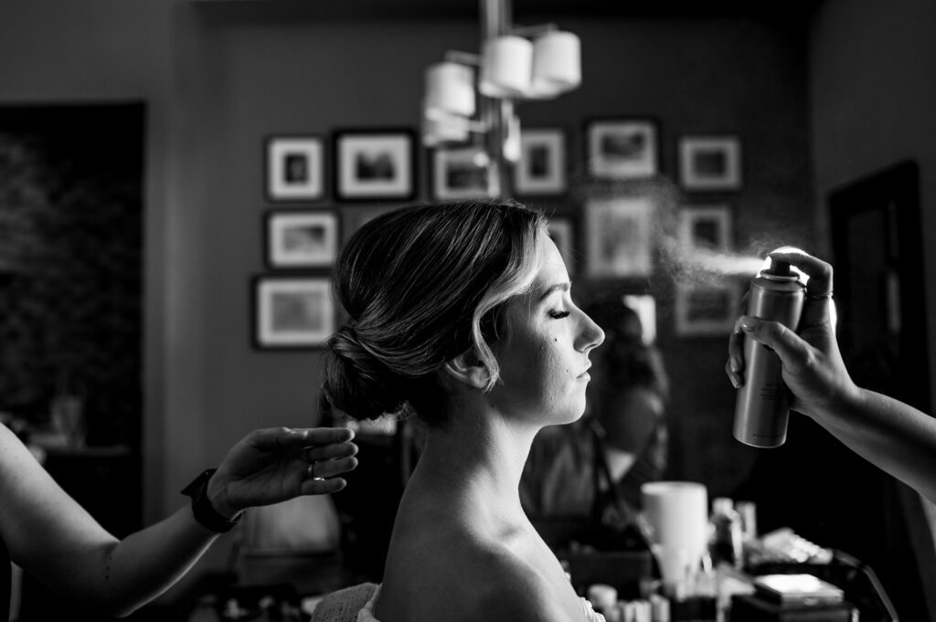 A black-and-white photograph of a bride getting ready on her wedding day. Sitting with eyes closed, one stylist sprays a fine mist of hairspray over exquisitely done-up hair, while another gently tugs at tresses to make sure everything is just so. The softly blurred background frames a few framed pictures on the wall, making this a cozy and intimate bridal preparation scene.