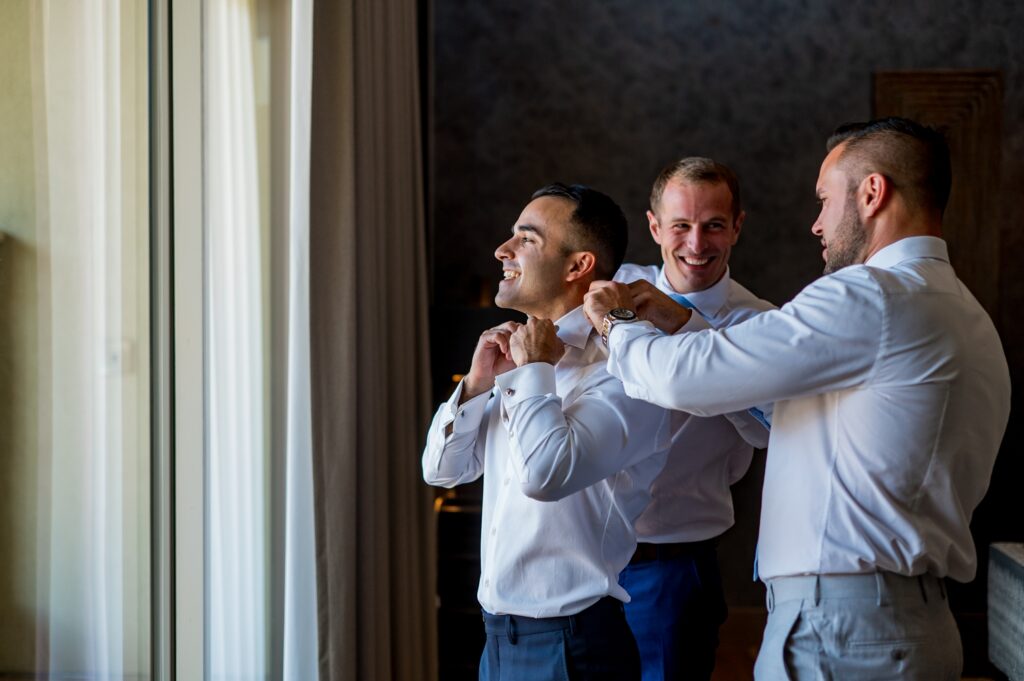 The groom gets ready for his wedding, smiling as one of his groomsmen adjusts his collar, with another looking on over his shoulder, beaming. The scene is naturally lit from a window nearby, showing the fun and camaraderie between the groom and his closest friends as they get ready for the big day.