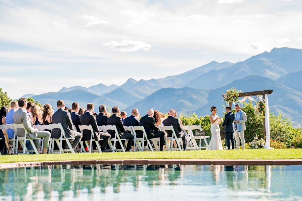 Garden of the Gods Resort-outdoor wedding ceremony, in which the bride and groom were exchanging vows before a flower-ornamented wooden arch. Guests were in neat rows of chairs, wearing formal attire and watching the couple with much interest. The stunning mountain backdrop, along with a clear blue sky, forms a picturesque setting; reflections of this ceremony in the pool add serenity and romance to the scene.