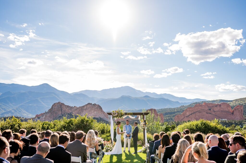 A stunning outdoor wedding ceremony at Garden of the Gods Resort: the bride and groom stand at a flowery, arched wooden structure, promising vows to each other as the sun shines brightly overhead. The guests sit in neat rows, and the backdrop is a breathtaking view of towering red rock formations and majestic peaks of the Rocky Mountains-a picturesque, romantic setting for the couple's special moment.