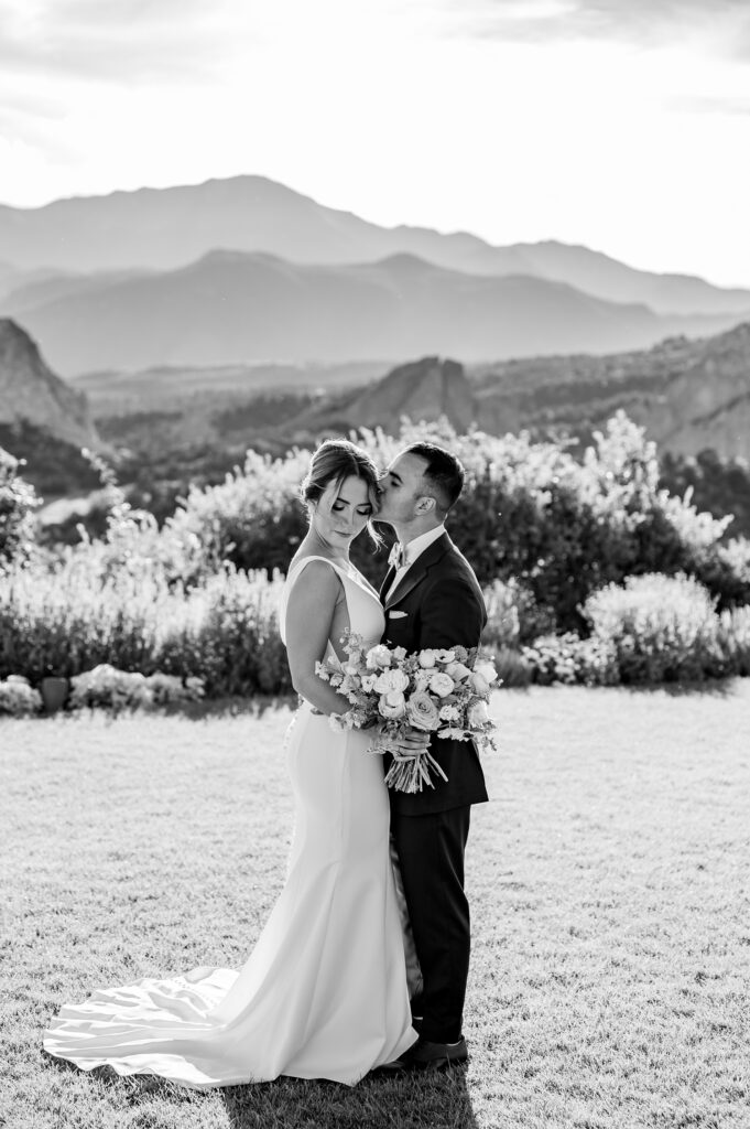 A black-and-white wedding portrait of a bride and groom in Colorado, taken at the Garden of the Gods Resort: The groom gently presses a kiss to his bride's temple while she looks down and clasps her rose and florist bouquet. The mountain landscape-a backdrop of rustic red rock formations-fills the frame, completing this classic, romantic scenario.