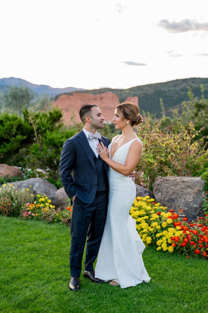 A newlywed couple stands in a lush garden at the Garden of the Gods Resort in Colorado Springs, accented with flowers of vibrant yellow and red. The bride in her sleek white gown rests her hand softly on the groom's chest as she looks into his eyes. The groom, attired in a classic navy suit with bow tie, looks lovingly at his bride. The red rock formations and rolling hills forming the backdrop add to the romantic and scenic view.
