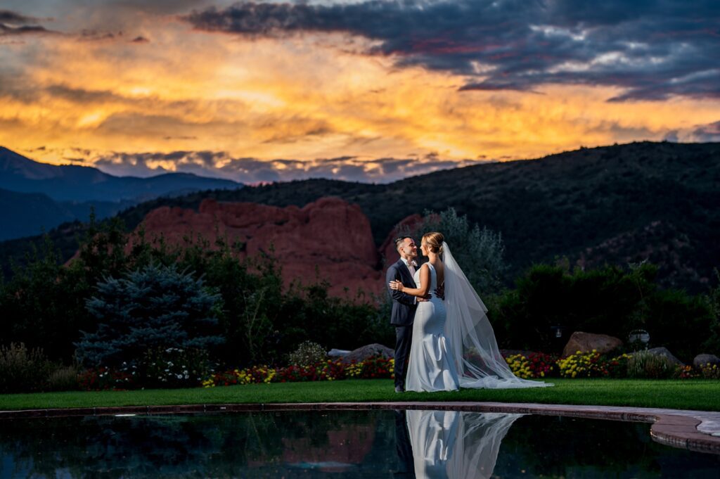 Wedding Portrait-Colorado Springs Garden of the Gods Resort Newlyweds make a stunning evening scene standing next to a reflective pool as a vibrant sunset transforms the sky overhead with shades of orange, yellows, and deep blues into the romance and warms the red rock formation backdrop. The bride, in a sleek white gown flowing with a veil, looks lovingly at her groom while their reflection in the water adds to the dreamy atmosphere.