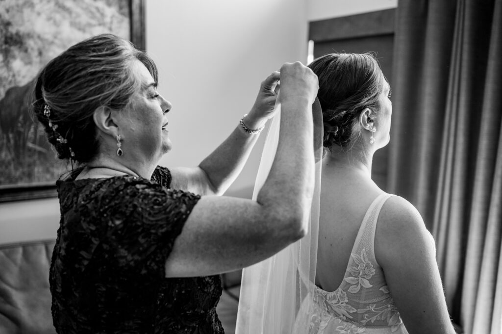 Feelings are at the forefront in this black and white picture taken during the Beaver Run Resort wedding ceremony. It depicts a mother clasping on a lace veil onto her daughter's bridal hair. Portrayed in the picture is the mother clad in a beaded gown dress and the daughter looking elegant in a lace wedding gown. They are in the midst of adjusting the veil in a well-lit room.