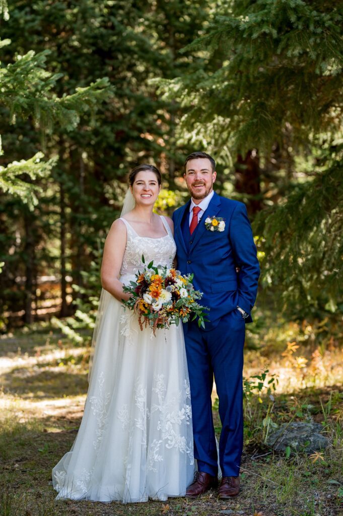 Bride and groom pose in a scenic forest setting before their wedding ceremony in Colorado. The bride who is wearing a lace wedding dress and veil holds the vibrant bouquet of autumn flowers, while the groom, in a navy blue suit with a red tie, smiles beside her. A beautiful outdoor wedding portrait surrounded by towering pine trees in the Colorado mountains.