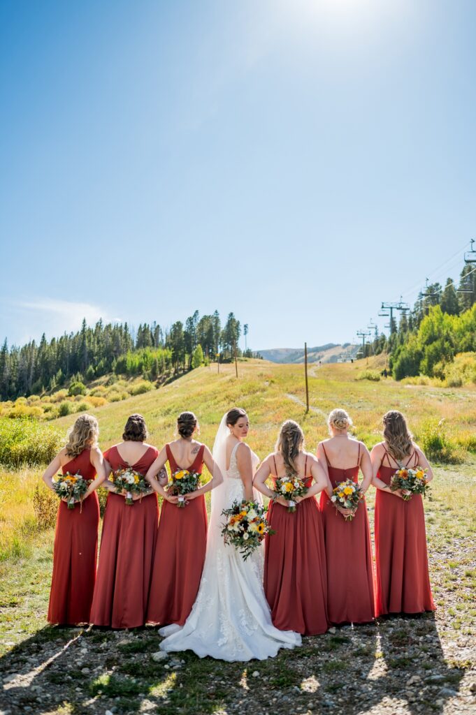 In a scenic Colorado mountain backdrop, the bride and bridesmaids in rust-colored dresses with bouquets pose. Photos of a beautiful alpine meadow and ski lift in the background were taken with a drone at the wedding held outside in the fall.
