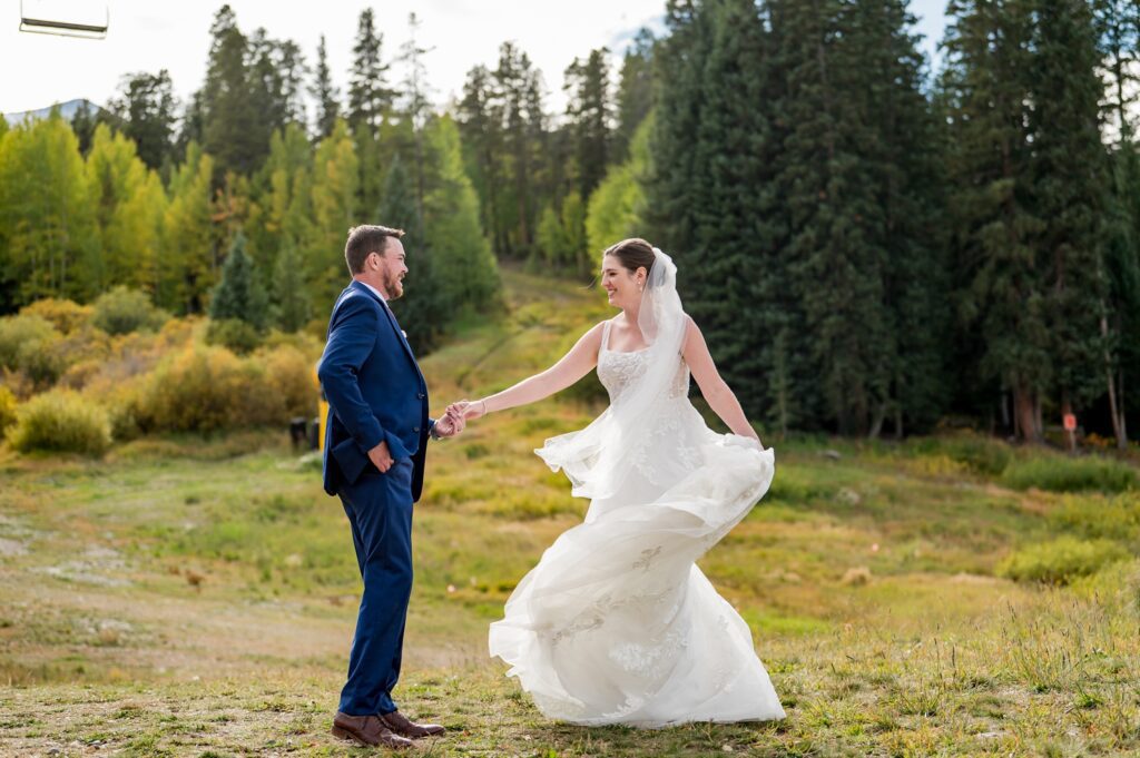 The bride twirled around in her wedding dress as she held her groom's hand at their Colorado mountain wedding venue. The outdoor fall wedding featuring a beautiful landscape of greenery and gold foliage along with the tall pine trees in the background. The photo was taken by a Colorado wedding photographer who is an expert in natural and romantic wedding photography.