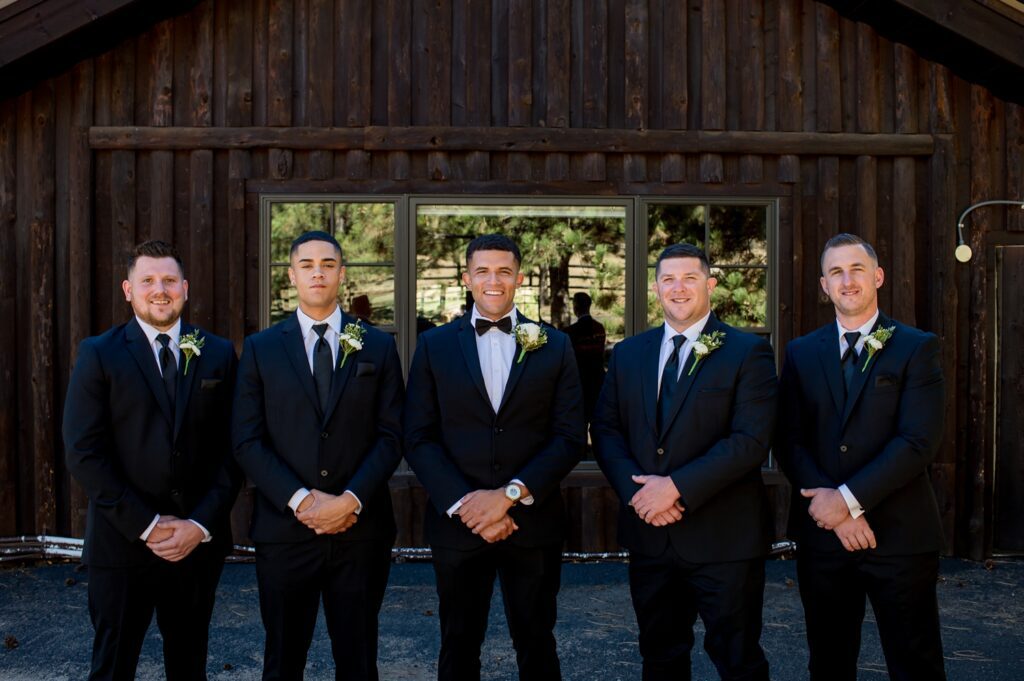 Classic black suits worn by groom and groomsmen posed outside the rustic wooden barn at Spruce Mountain Ranch in Colorado. True-to-color wedding photography capturing the groom's party at this stunning Colorado wedding venue.