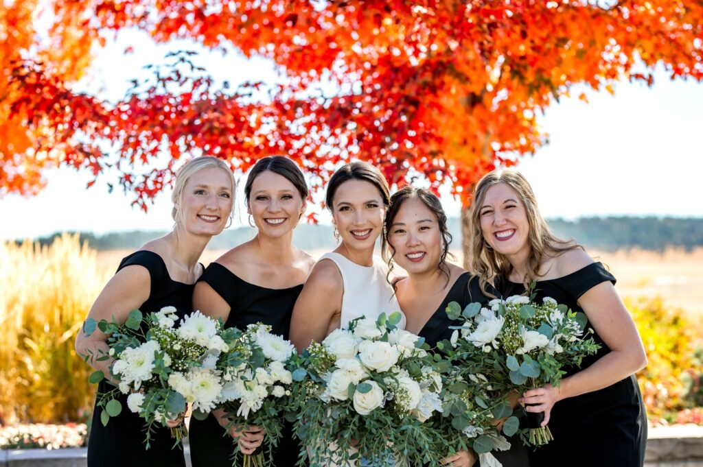 Bride and bridesmaids pose with elegant white bouquets in front of vibrant red fall foliage at Spruce Mountain Ranch in Colorado. True-to-color wedding photography capturing the beauty of an autumn wedding at one of Colorado's premier wedding venues.