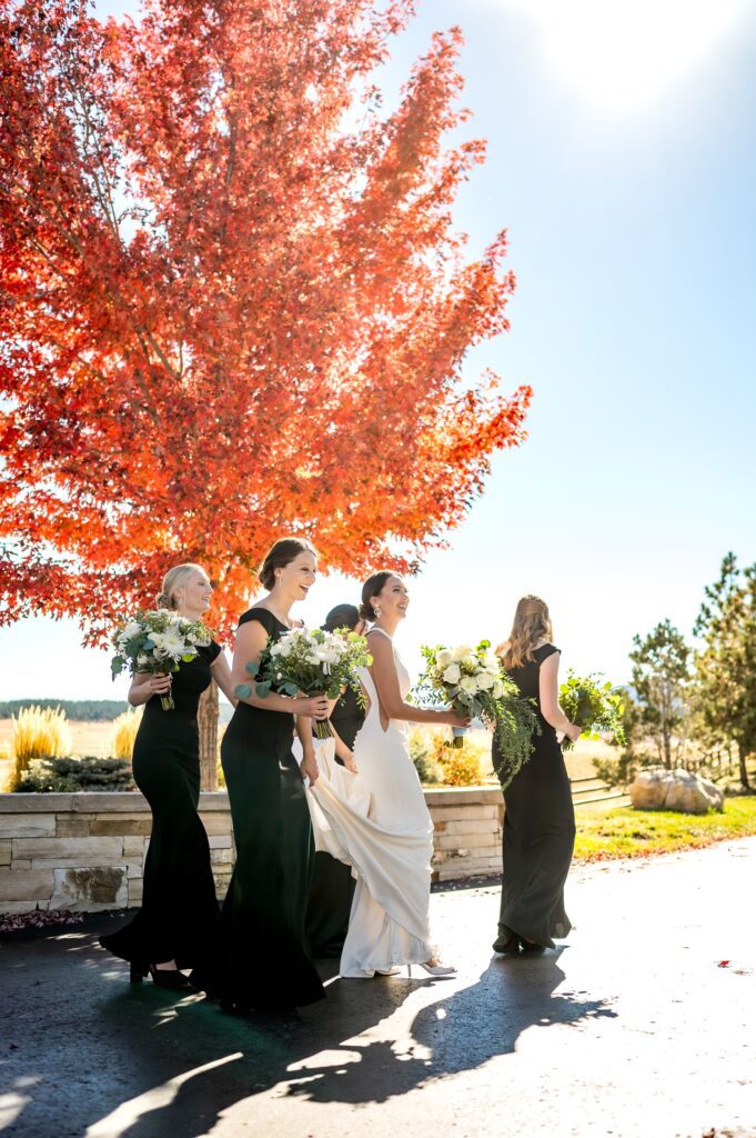 Bride and bridesmaids walking with white floral bouquets under the vibrant red fall foliage of Spruce Mountain Ranch in Colorado. True-to-color wedding photography capturing an elegant fall wedding at a top Colorado wedding venue.