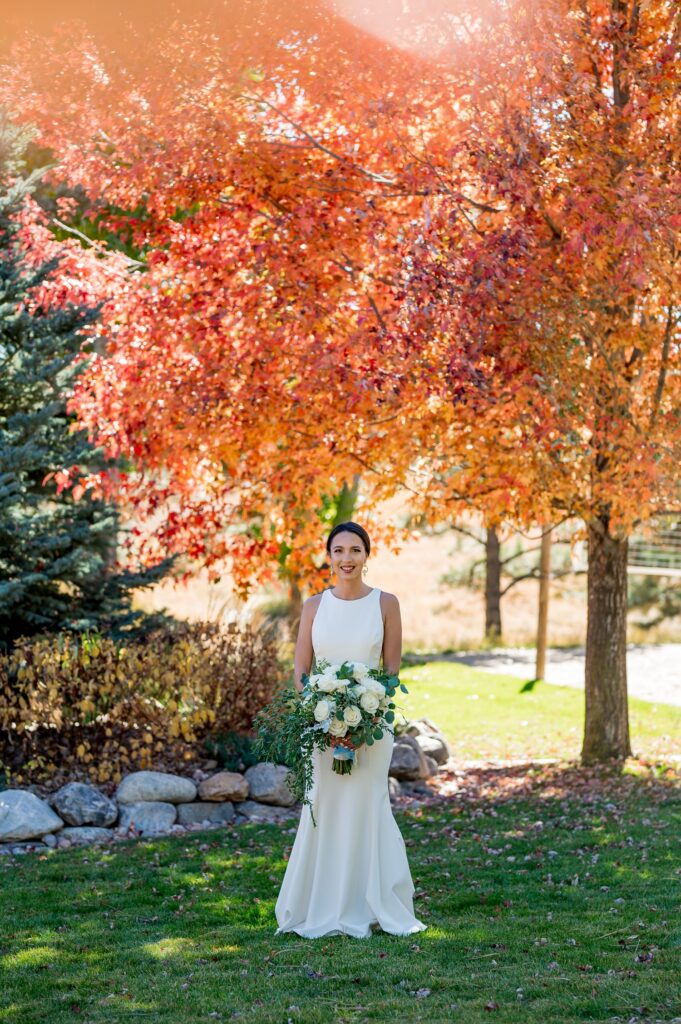 The bride, with a traditional white bouquet, sits among the bright red and orange of the fall foliage at Spruce Mountain Ranch, Colorado. True to color wedding photography to capture this beautiful autumn wedding at one of Colorado's premiere wedding venues.