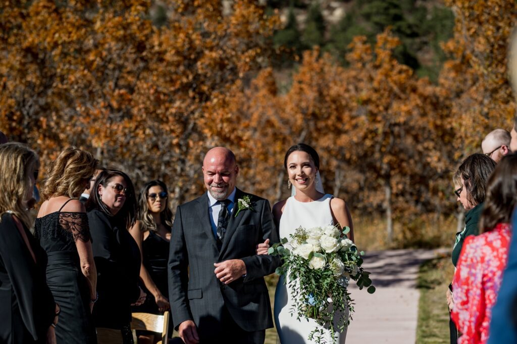 The bride walks down the aisle at this stunning Spruce Mountain Ranch fall wedding with her father, golden fall leaves, and stunning wedding guests joining in framing such a sweet special moment taken by a true to color Colorado Wedding Photographer.