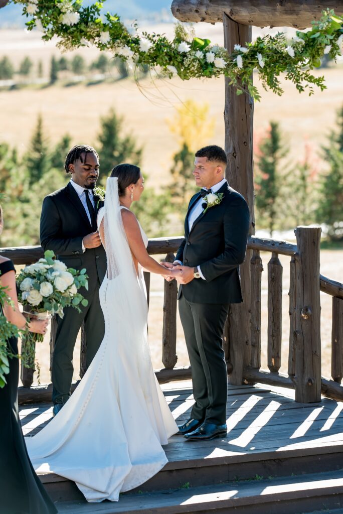 Bride and groom exchange vows under a floral-adorned wooden arbor at their outdoor wedding ceremony at Spruce Mountain Ranch in Colorado. True-to-color wedding photography capturing a heartfelt moment at a top Colorado wedding venue.