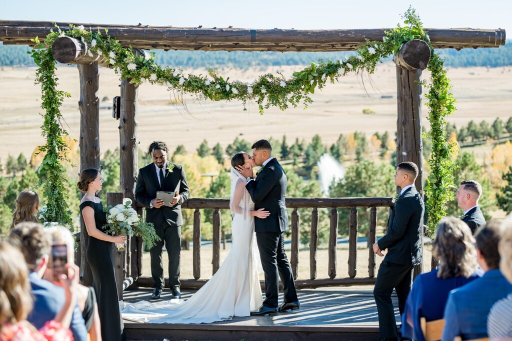 Bride and groom share their first kiss as husband and wife under a floral-adorned wooden arbor at Spruce Mountain Ranch in Colorado. True-to-color wedding photography captures this romantic outdoor ceremony with breathtaking fall scenery.