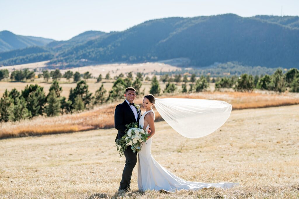 The bride and groom of Spruce Mountain Ranch are in the open field with the bride's veil blowing beautifully in the wind. Breathtaking Colorado fall wedding captured with true-to-color photography to showcase this stunning mountain venue.